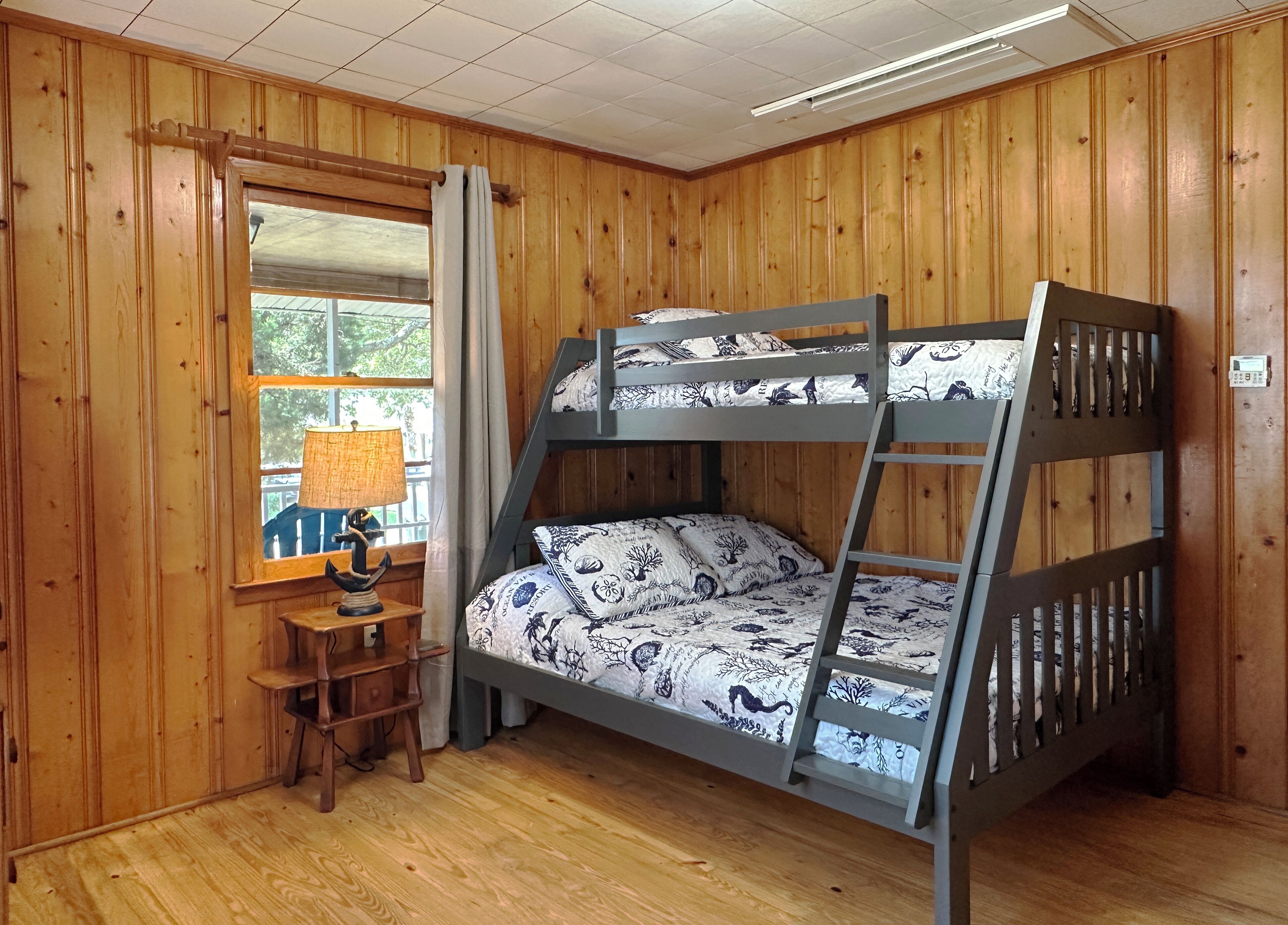 A bunk bed with patterned bedding, a wooden desk with a lamp, and a window with curtains.