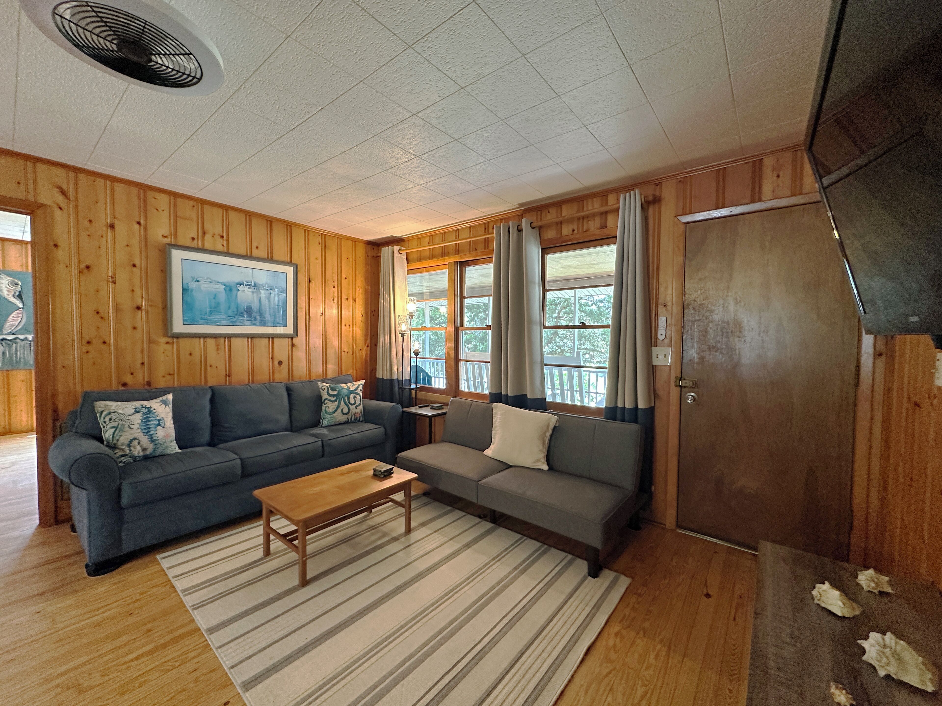 A cozy cabin interior with wooden walls, a gray sofa, a striped rug, and a wooden table.