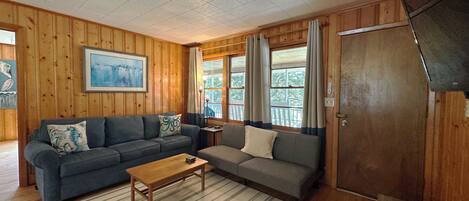 A cozy cabin interior with wooden walls, a gray sofa, a striped rug, and a wooden table.