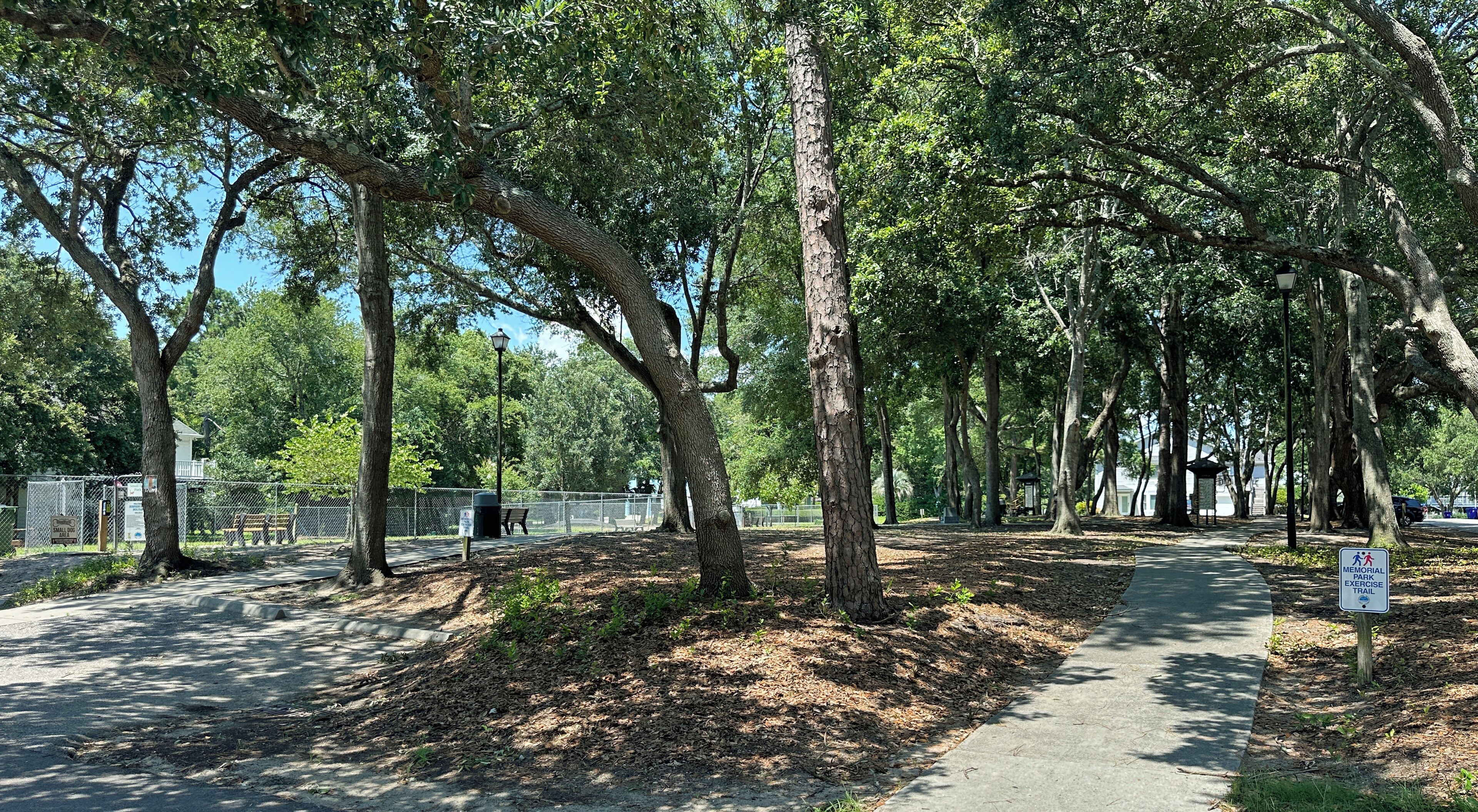 A park with a paved walkway, trees, benches, and a memorial exercise sign.