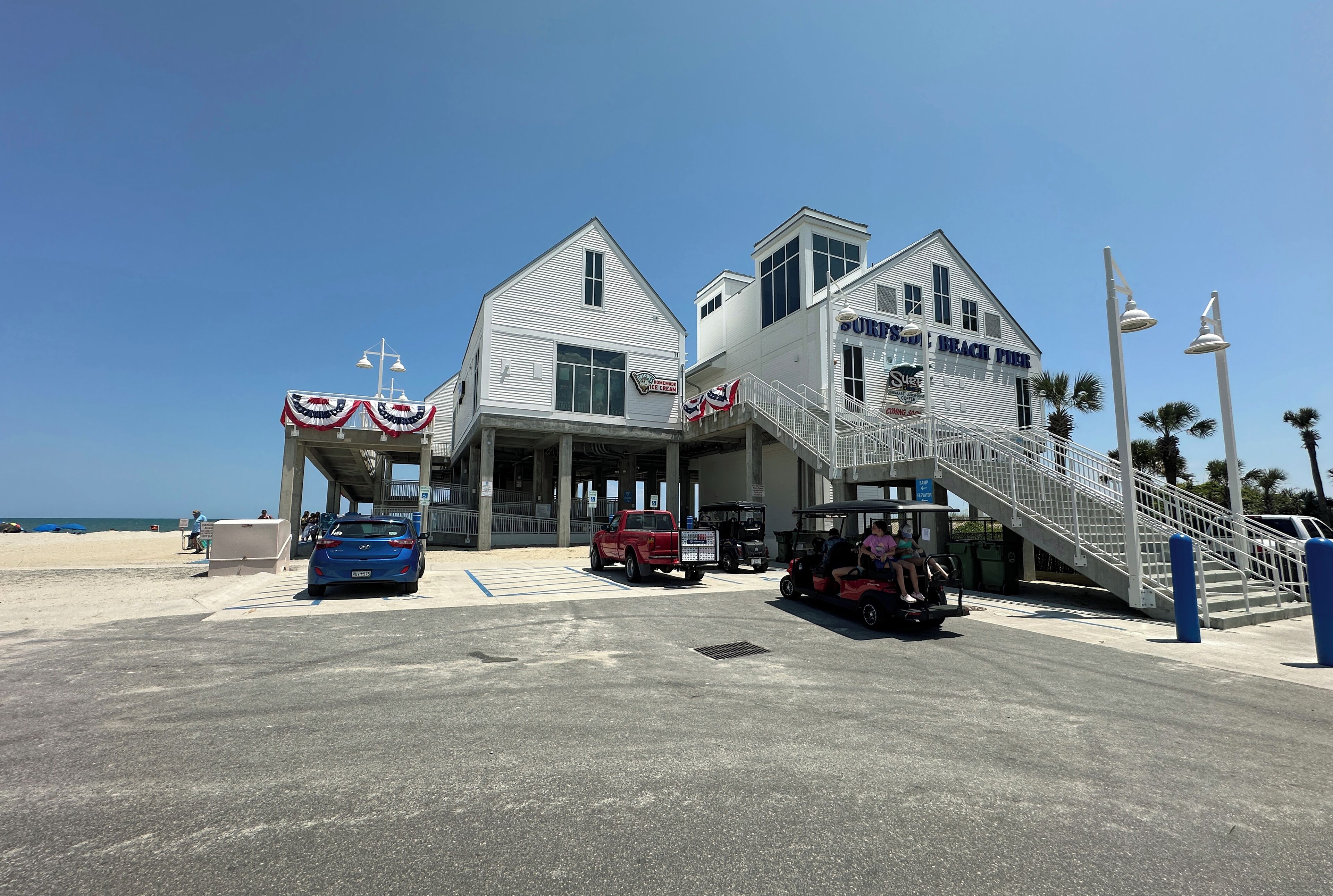 A beachside building with a staircase leading to a beach entrance, a sign for 'Surf's Up Beach Pier', and a red truck parked in front.