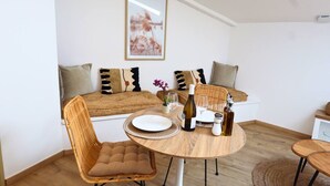 A cozy room with a round wooden table, wicker chairs, a sofa with patterned cushions, and a framed artwork on the wall.