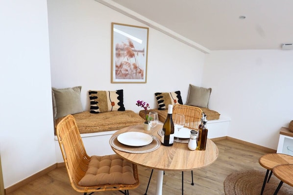 A cozy room with a round wooden table, wicker chairs, a sofa with patterned cushions, and a framed artwork on the wall.