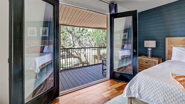 A bedroom with a wooden floor, a bed with a white quilt, a nightstand with a lamp, and a view of a balcony with a railing. - Florida Breeze |Pool & Jacuzzi| 2-min walk to BCH (St. Augustine)