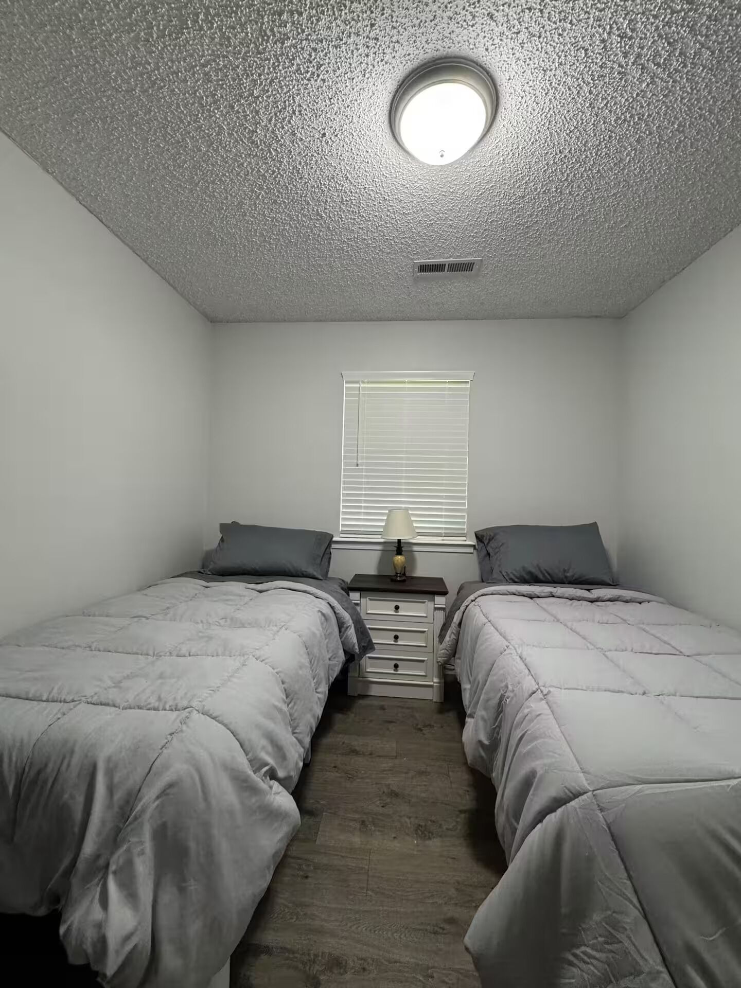 Ein Schlafzimmer mit zwei Einzelbetten, einem Nachttisch mit Lampe, einem Fenster mit Jalousien und einer strukturierten Decke.