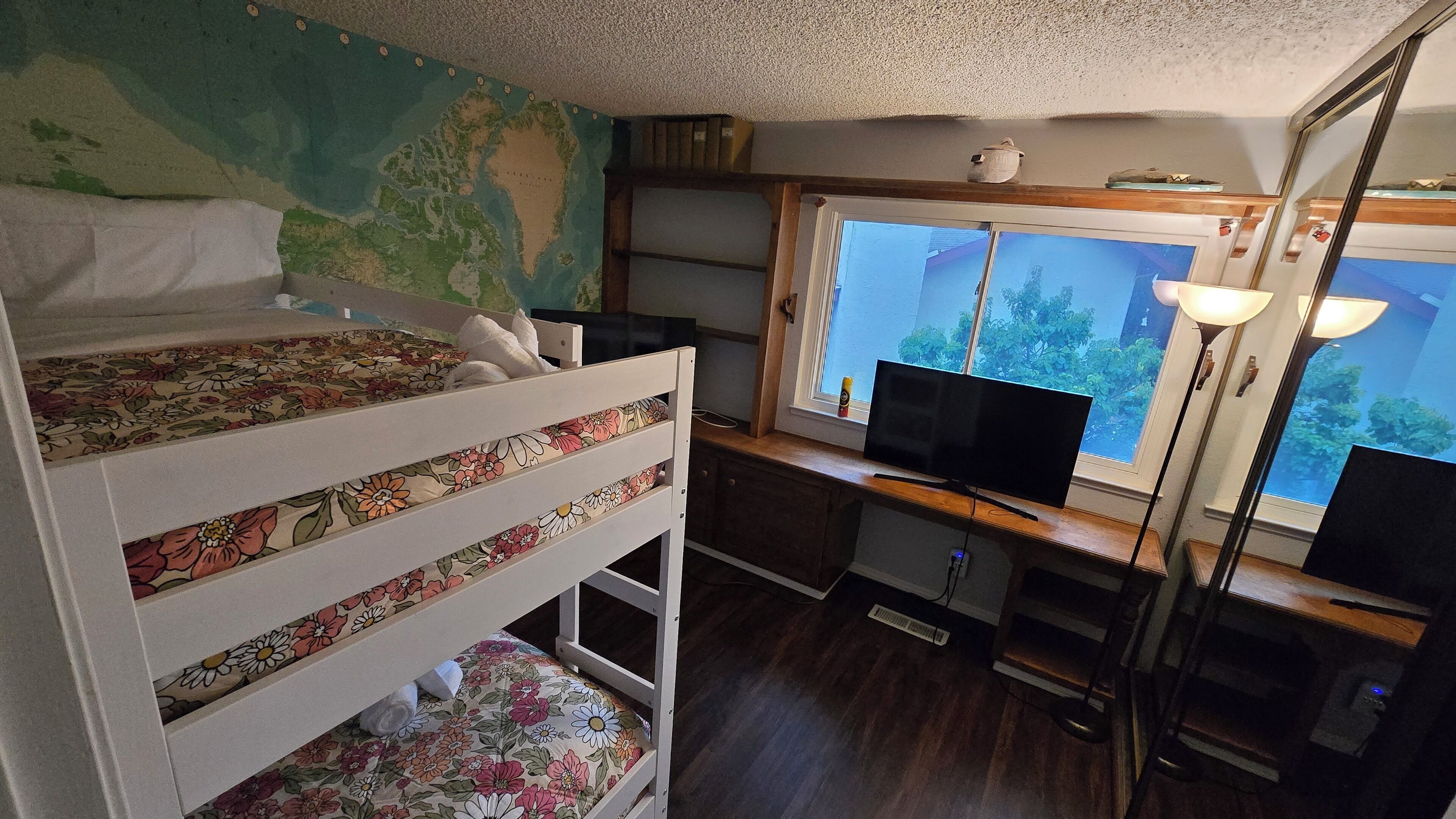Una cama individual con ropa de cama estampada, un escritorio con computadora y lámpara, y una ventana que da a vistas de vegetación.