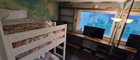 Una cama individual con ropa de cama estampada, un escritorio con computadora y lámpara, y una ventana que da a vistas de vegetación.