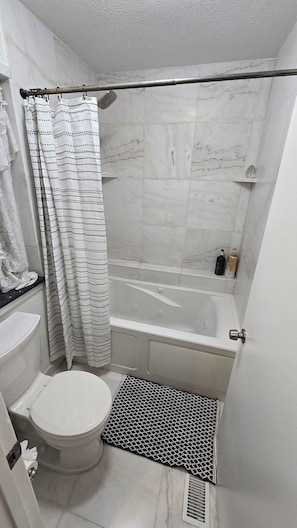 A compact bathroom with a toilet, a shower with a curtain, a bathtub, and a small shelf with toiletries.
