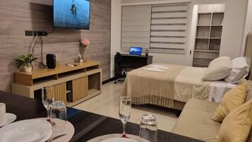 Una habitación de hotel moderna con televisor de pantalla plana, una unidad de entretenimiento de madera, una cama con manta color beige y un área de comedor con mesa dispuesta para dos.