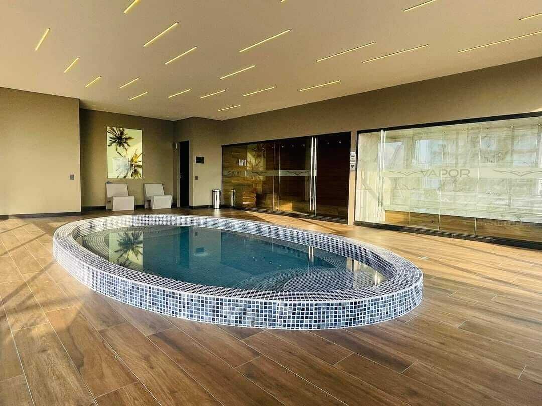 Une piscine intérieure moderne, avec un bord carrelé, entourée d’un plancher en bois et de murs en verre.