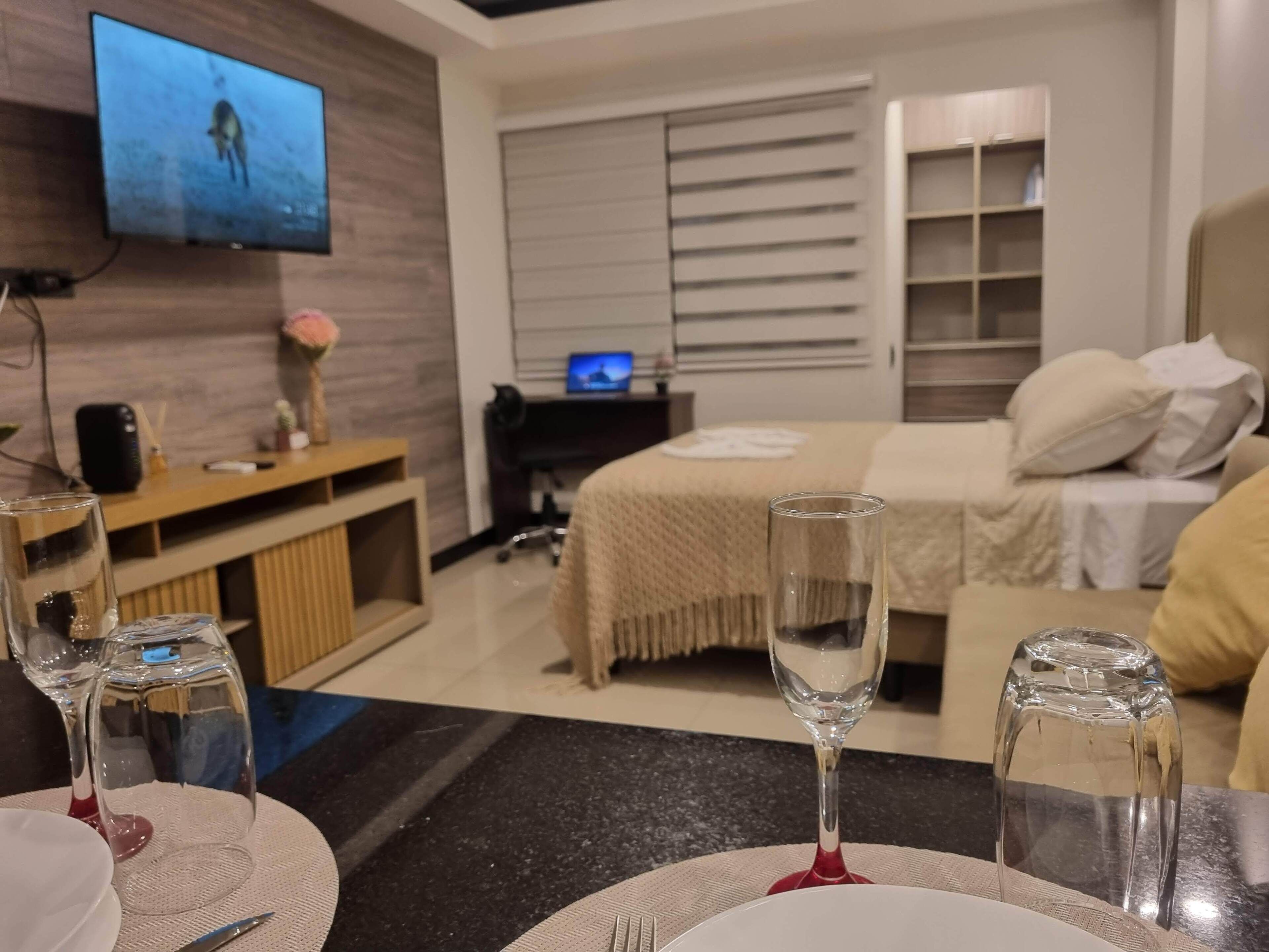 Habitación de hotel con una cama, un televisor montado en la pared, un escritorio con una computadora y un área de comedor con mesa para dos.