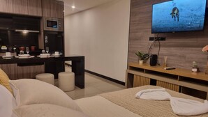 Une chambre d’hôtel moderne équipée d’un lit, d’un téléviseur fixé au mur, d’un petit coin cuisine et d’un espace salon avec des tabourets.