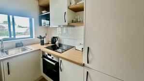 A modern kitchen with white cabinets, a black oven, a stainless steel sink, and a window with a view of greenery. - AZORES PEDRA APARTMENTS - G (Fajã de Baixo)