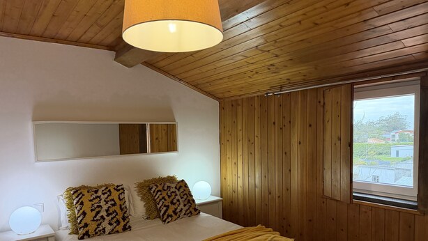 Une chambre avec un plafond en bois, un lit avec une literie blanche et des oreillers à motifs, une lampe de chevet, un miroir et une fenêtre donnant sur des immeubles.