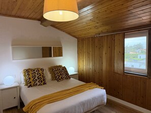 Une chambre avec un plafond en bois, un lit avec une literie blanche et des oreillers à motifs, une lampe de chevet, un miroir et une fenêtre donnant sur des immeubles.