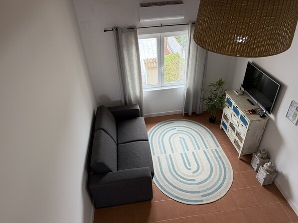 Un salon avec un canapé, un téléviseur posé sur un meuble en bois, un grand luminaire en fibres tressées et une vue sur l’extérieur par une fenêtre ornée de rideaux.