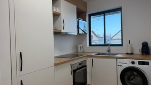 A compact kitchen with white cabinets, a built-in oven, a washing machine, and a window with a view of a building. - AZORES PEDRA APARTMENTS A (Fajã de Baixo)