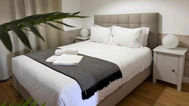 A neatly made bed with white linens, a grey headboard, and a black and white throw blanket. There is a white bedside table with a lamp and a small round table beside the bed. - AZORES PEDRA APARTMENTS A (Fajã de Baixo)