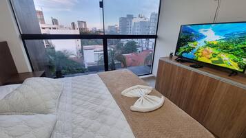 A bedroom with a large bed, a wooden dresser, a television, and a view of the city through a large window.
