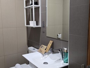 A modern bathroom with a white sink, a mirror, and a shelf above the toilet holding toiletries.