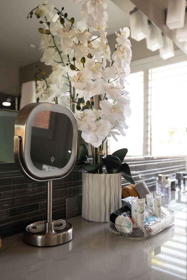 Un baño con un espejo grande, orquídeas blancas, artículos de aseo y un perchero para toallas.
