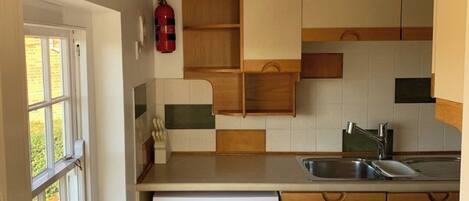 Une cuisine compacte avec des meubles blancs, un lave-vaisselle encastré, une étagère en bois et une fenêtre donnant sur un espace verdoyant.