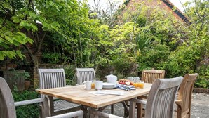 A wooden table set outdoors with chairs, surrounded by lush greenery and a brick building in the background.