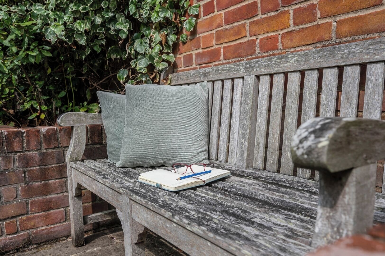 A wooden bench with a cushion and a notebook with glasses and a pencil on it.