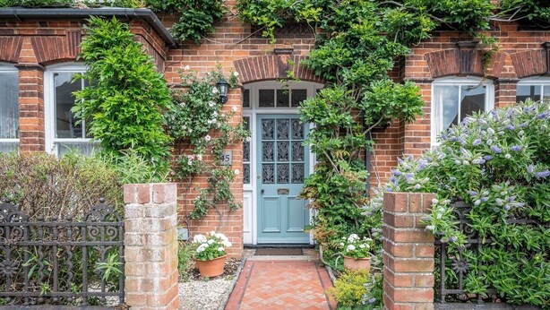 A brick house with a blue door, surrounded by lush greenery and a brick pathway leading to the entrance.