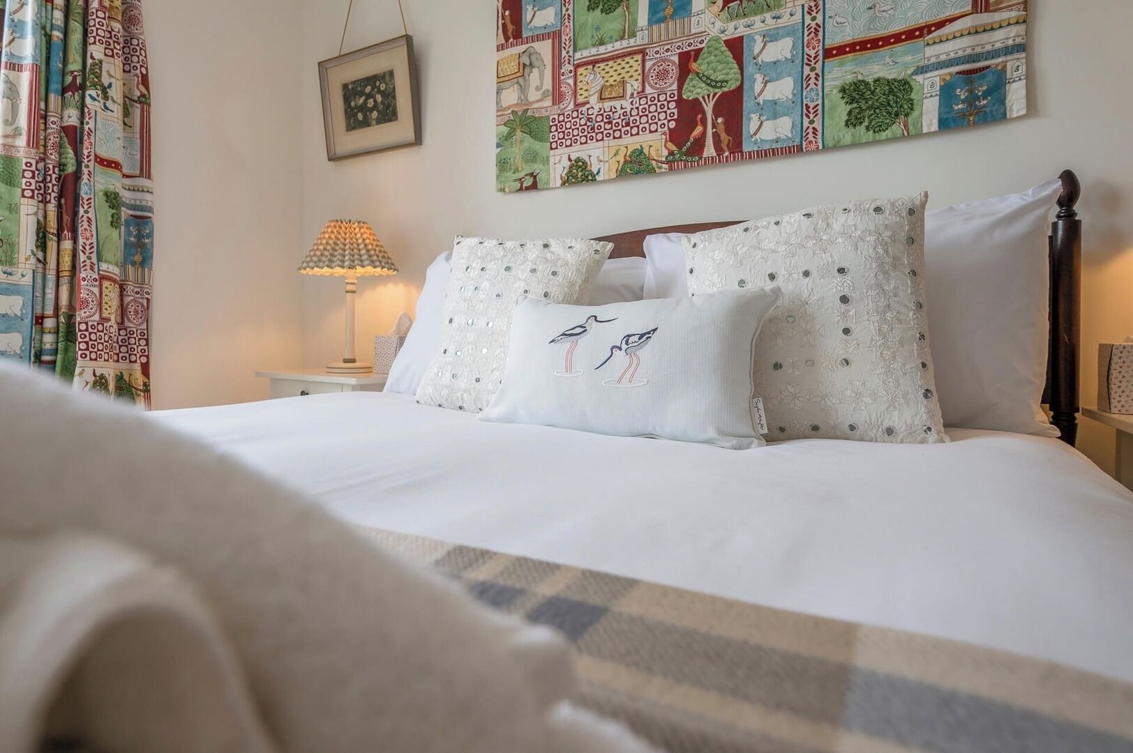 A neatly made bed with white linens and decorative pillows, a bedside lamp, and a colorful tapestry on the wall.