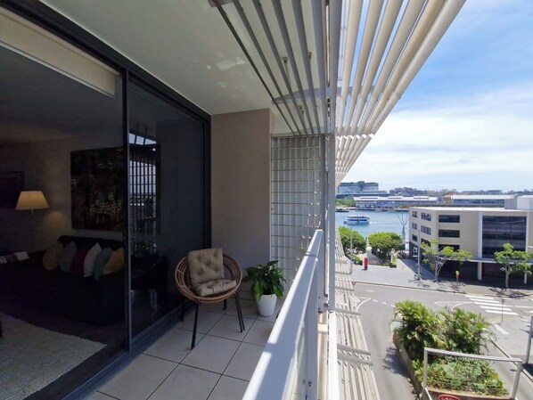 A balcony with a view of a cityscape and water. There is a chair and a potted plant on the balcony. - KozyGuru | Barangaroo | Waterfront Views Studio (Sydney)