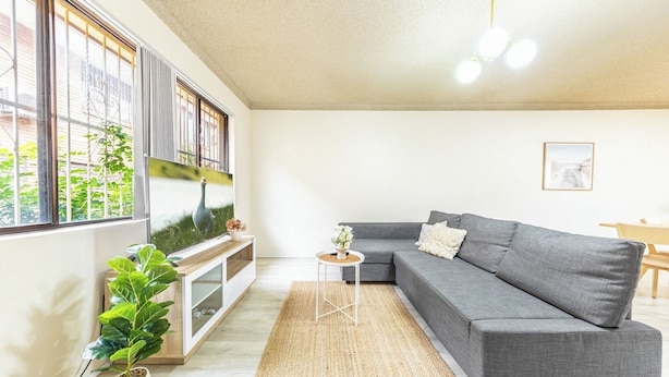 A living room with a grey sofa, a TV on a white stand, a small round table with a vase, and a large window with blinds. - KozyGuru | Cabramatta | Spacious 3BR Townhouse (Cabramatta)