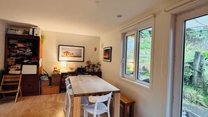 A room with a wooden dining table, white chairs, a wooden cabinet with various items, a framed picture on the wall, and a window with a view of greenery outside. - 3 Bedroom Passive Haus in the lush Aro Valley (Wellington)
