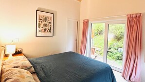 A bedroom with a bed, a nightstand, a lamp, a framed botanical print, and a window with curtains. - 3 Bedroom Passive Haus in the lush Aro Valley (Wellington)