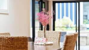 Una mesa de comedor con un florero de flores rosas, sillas de mimbre y vistas al exterior a través de amplios ventanales.