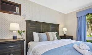 A hotel room with a bed, two bedside tables with lamps, a wooden headboard, and a window with curtains.
