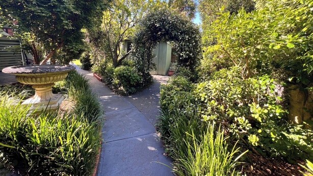 Ein Gartenweg mit einer Steinbrunnen, umgeben von üppigem Grün und einem hölzernen Tor.