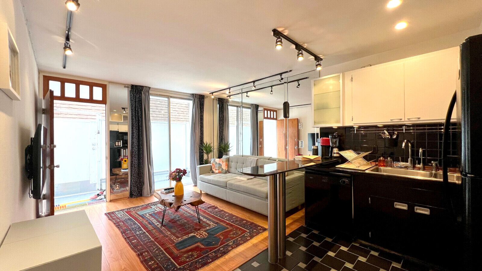 Una cocina moderna con un refrigerador negro, un horno empotrado y un lavamanos. Hay un comedor con mesa y sillas, y una sala de estar con un sofá y una mesita de centro.