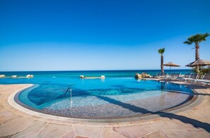 A clear blue pool with a curved edge, surrounded by lounge chairs and palm trees, overlooking the ocean.