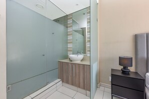 A modern bathroom with a glass shower enclosure, a wooden vanity with a sink, and a bedside table with a lamp.