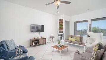 Ein modernes Wohnzimmer mit Deckenventilator, Flachbildfernseher, Sofa, Couchtisch und Fenster mit Blick.
