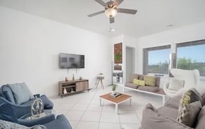 A modern living room with a ceiling fan, a flat-screen TV, a sofa, a coffee table, and a window with a view. - Spacious 4-bedroom house with WiFi, fitness room in adorable Buckeye (Buckeye)