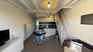Een moderne keuken met een centraal eiland, een eethoek met barkrukken, een trap en een woongedeelte met een bank en een televisie.