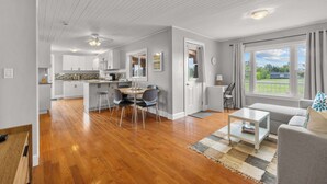 A spacious living area with a dining table, grey sofa, and a view of the outdoors. - Charming 2-Bed Cottage in Savage Harbour with Beach & Enclosed Sunroom (Prince Edward Island)