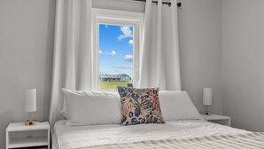 A bedroom with a large bed, white bedding, a colorful pillow, and a window with curtains. - Charming 2-Bed Cottage in Savage Harbour with Beach & Enclosed Sunroom (Prince Edward Island)