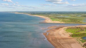 A coastal landscape with a sandy beach, a rocky breakwater, and a distant shoreline with buildings. - Charming 2-Bed Cottage in Savage Harbour with Beach & Enclosed Sunroom (Prince Edward Island)