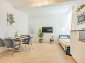 A modern, well-lit room with a dining area, a bed, a TV, and a kitchenette.
