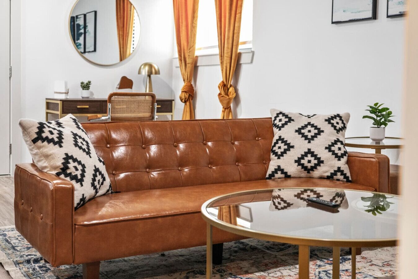 A brown leather sofa with patterned cushions, a round glass coffee table, a mirror, and a small side table with a plant.