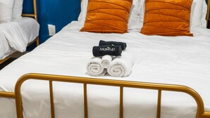 Habitación de hotel con cama, ropa de cama blanca, dos almohadas naranjas y una toalla negra con la palabra 'Makeup' escrita.