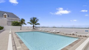 Une piscine rectangulaire à l’eau d’un bleu cristallin, entourée de chaises longues blanches et de palmiers, avec vue sur l’océan et un bâtiment en arrière-plan.